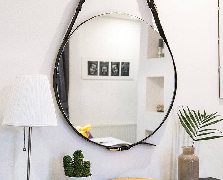 Un miroir rond à cadre noir, suspendu par une lanière de cuir, est placé au-dessus d'une table où se trouvent une lampe, un cactus et une plante.