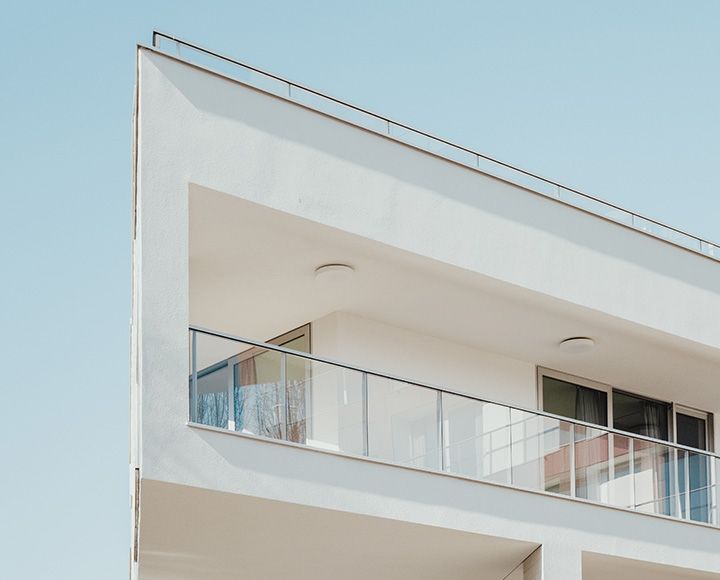 Vue en contre-plongée d'un angle de bâtiment moderne aux murs blancs, avec un balcon à balustrade en verre et un ciel bleu dégagé.