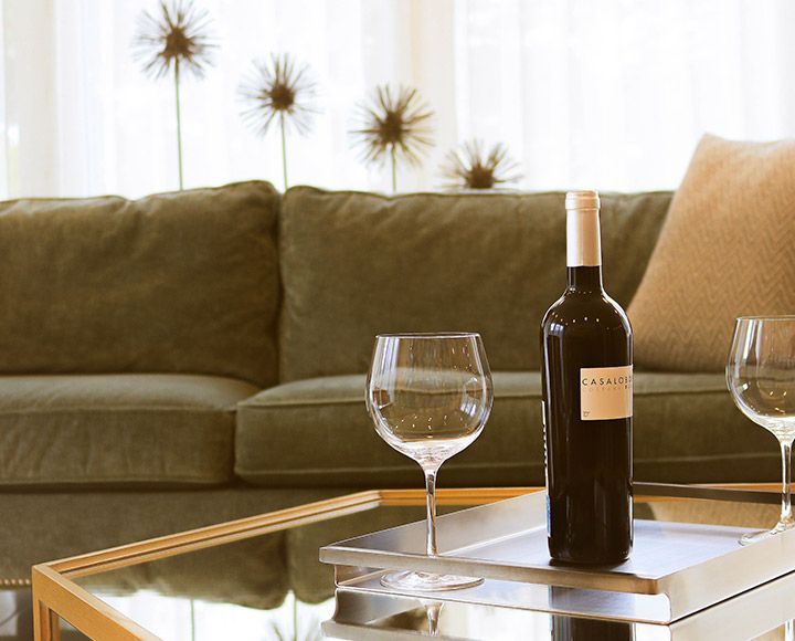 Une bouteille de vin et deux verres vides sur un plateau en argent posé sur une table basse en verre, avec un canapé vert en arrière-plan.