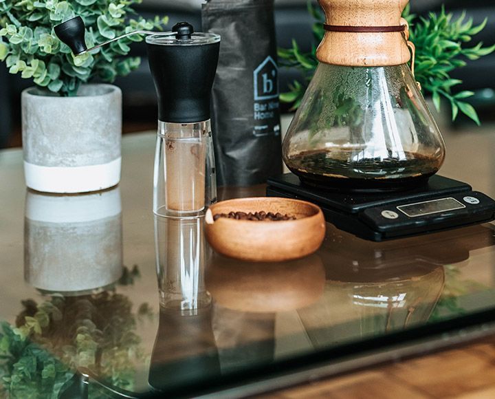Sur une table en verre, un nécessaire à café comprend une carafe Chemex posée sur une balance, un moulin à café manuel, un sachet de café et un bol de grains.