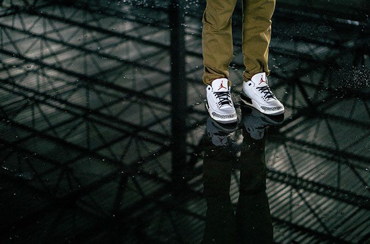 Une personne vêtue d'un pantalon beige et de baskets Jordan 3 blanches se tient au-dessus d'une surface sombre et réfléchissante à motif quadrillé.