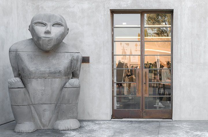 Une grande statue en pierre grise représentant une figure stylisée accroupie se trouve à côté d'une double porte vitrée dans un mur en béton.