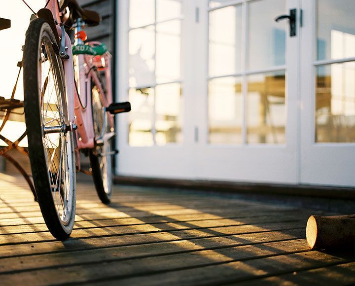 Un vélo rose est posé sur une terrasse en bois devant des portes vitrées au coucher du soleil, projetant de longues ombres.
