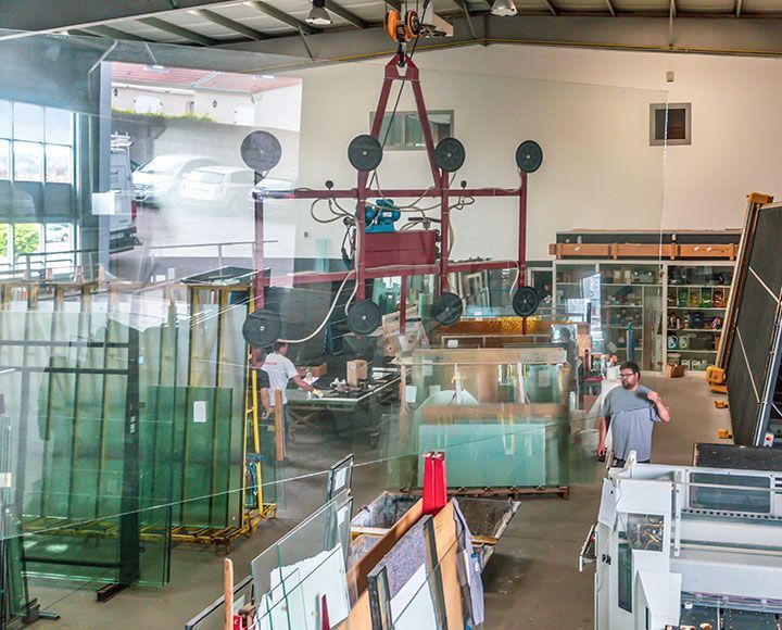 Un atelier de verrerie avec un grand cadre de levage par le vide suspendu, des feuilles de verre sur des étagères et des employés au travail.