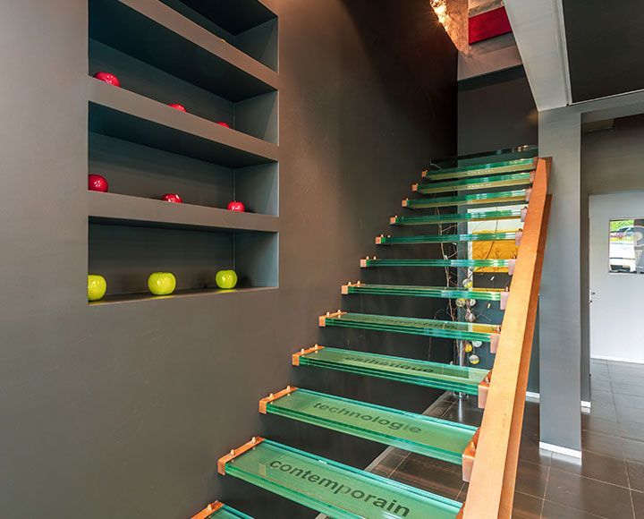 Un escalier aux marches en verre et à la rampe en bois, avec des murs sombres et des étagères présentant des pommes rouges et vertes.