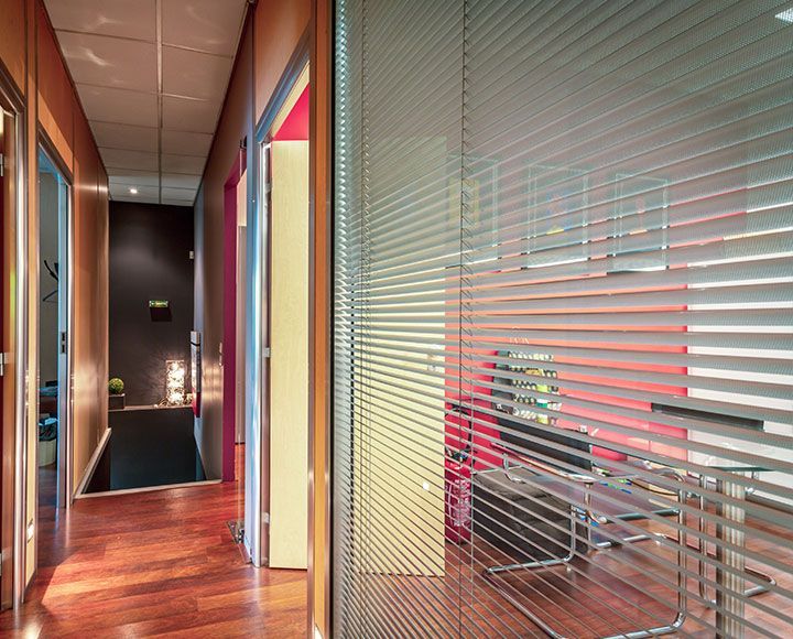 Un couloir au parquet mène à une pièce sombre, jouxtant une paroi de bureau vitrée recouverte de stores horizontaux.