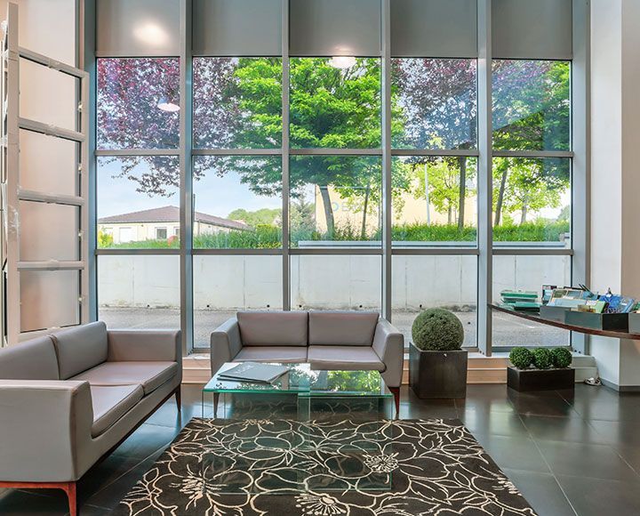 Un espace d'attente moderne avec deux canapés gris, une table basse en verre, un tapis à motifs et des baies vitrées.