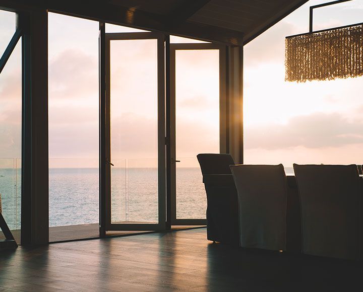 Salle à manger donnant sur l'océan à travers des portes vitrées ouvertes au coucher du soleil, avec un luminaire suspendu à franges.