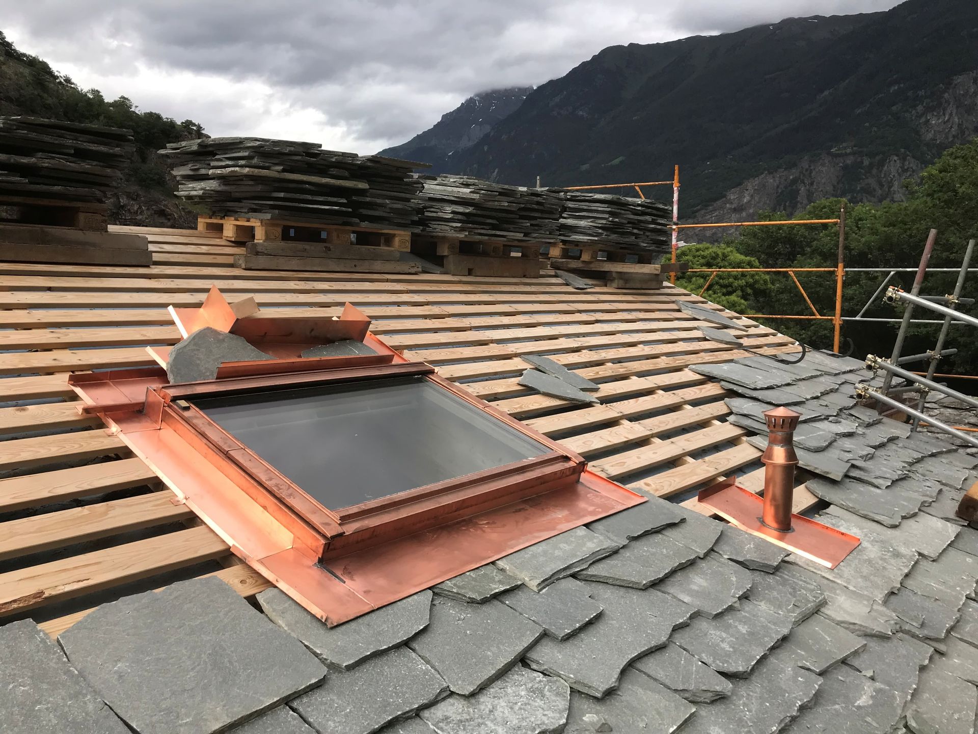 Toiture en construction avec fenêtre de toit, tuiles d'ardoise et matériaux de couverture empilés ; arrière-plan montagneux.