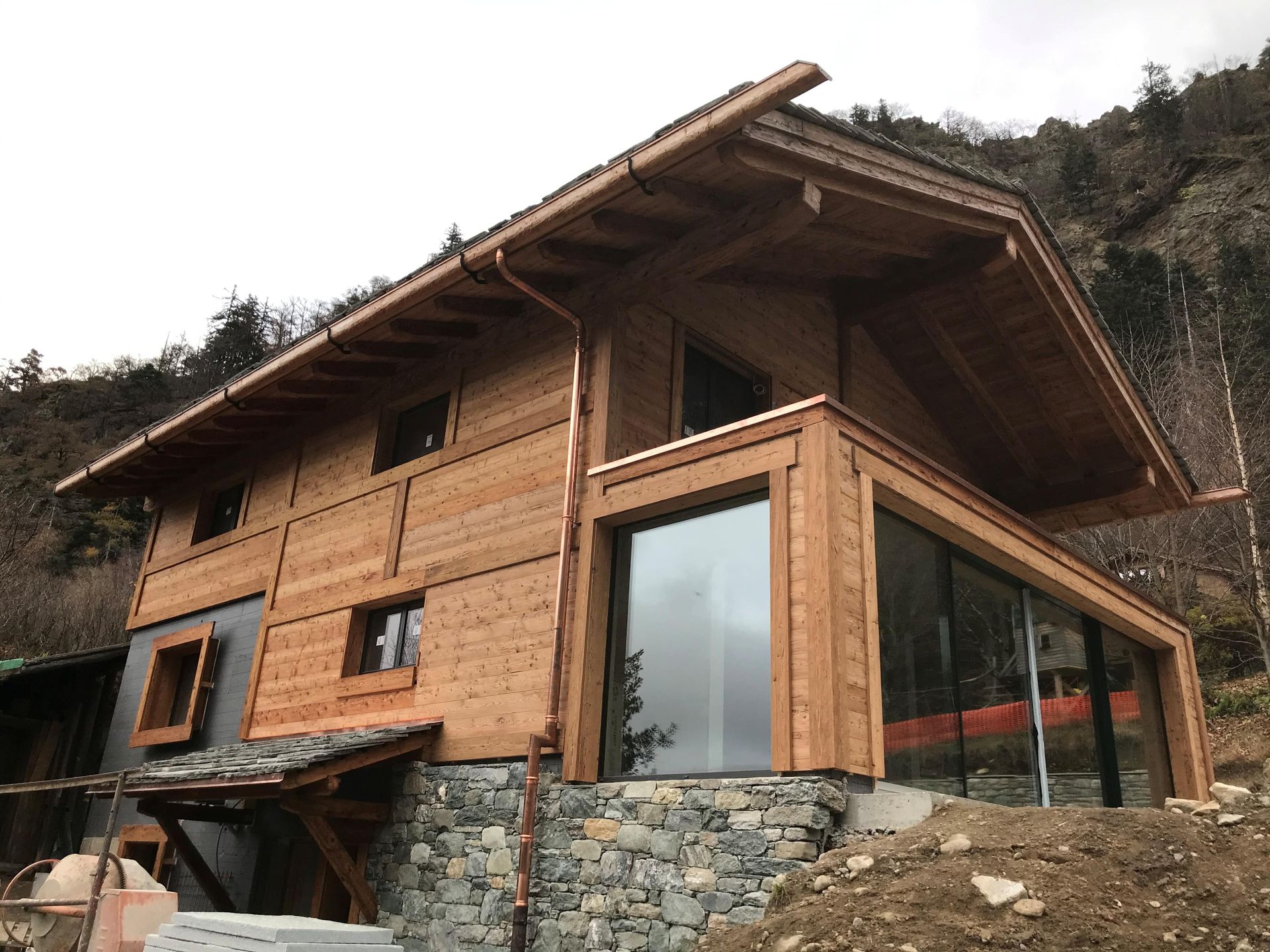 Maison de style chalet en bois, avec soubassement en pierre et grandes baies vitrées. Située à flanc de colline.