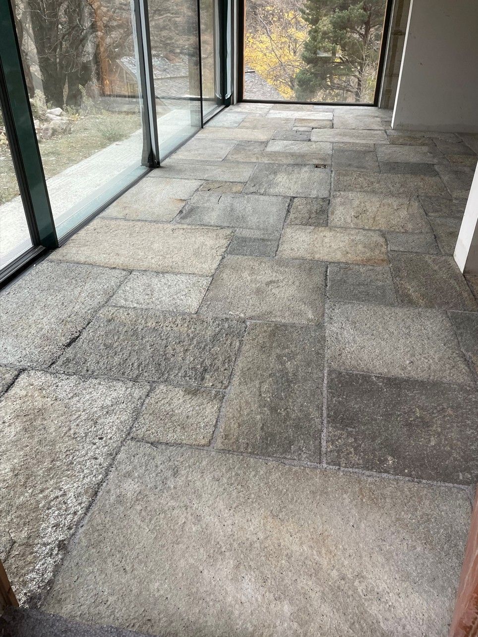 Sol carrelé en pierre dans une pièce baignée de soleil avec de grandes fenêtres donnant sur une zone boisée.