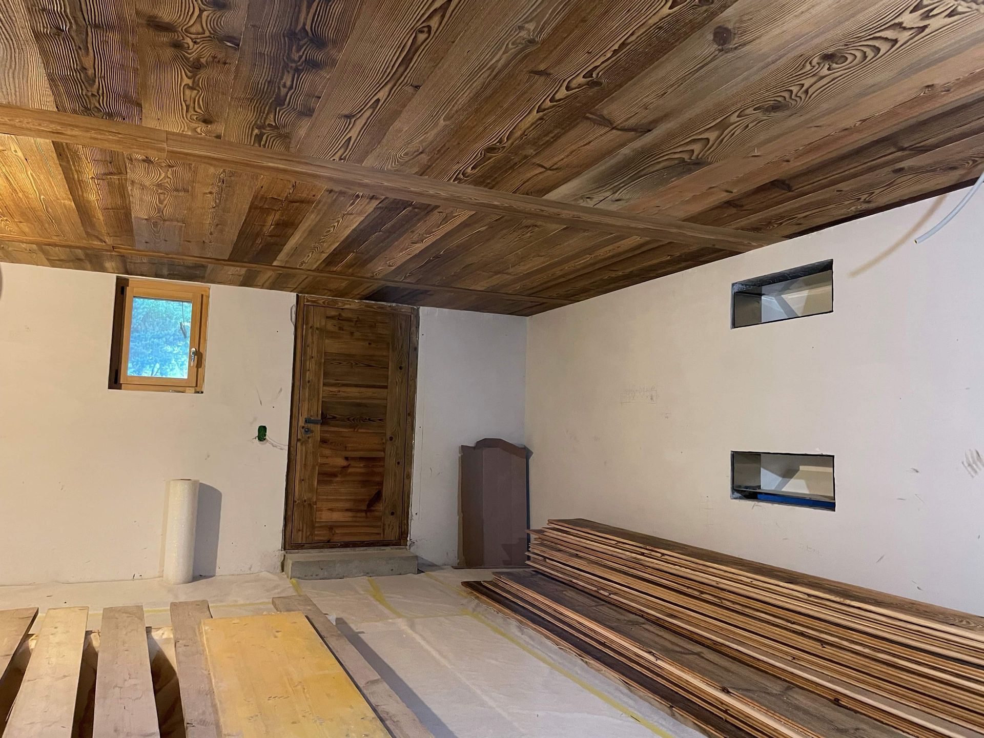 Intérieur d'une pièce avec plafond en bois, porte, fenêtre et plancher en planches de bois empilées.