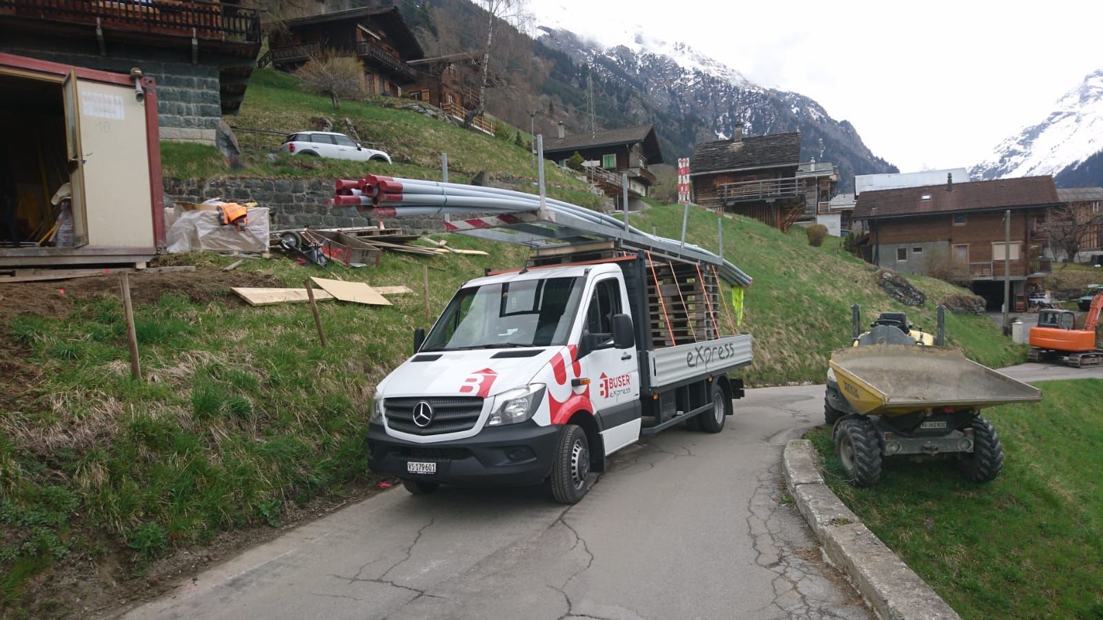 Un camion Mercedes blanc chargé de poutres métalliques circule sur une route à flanc de colline, près de bâtiments en bois.