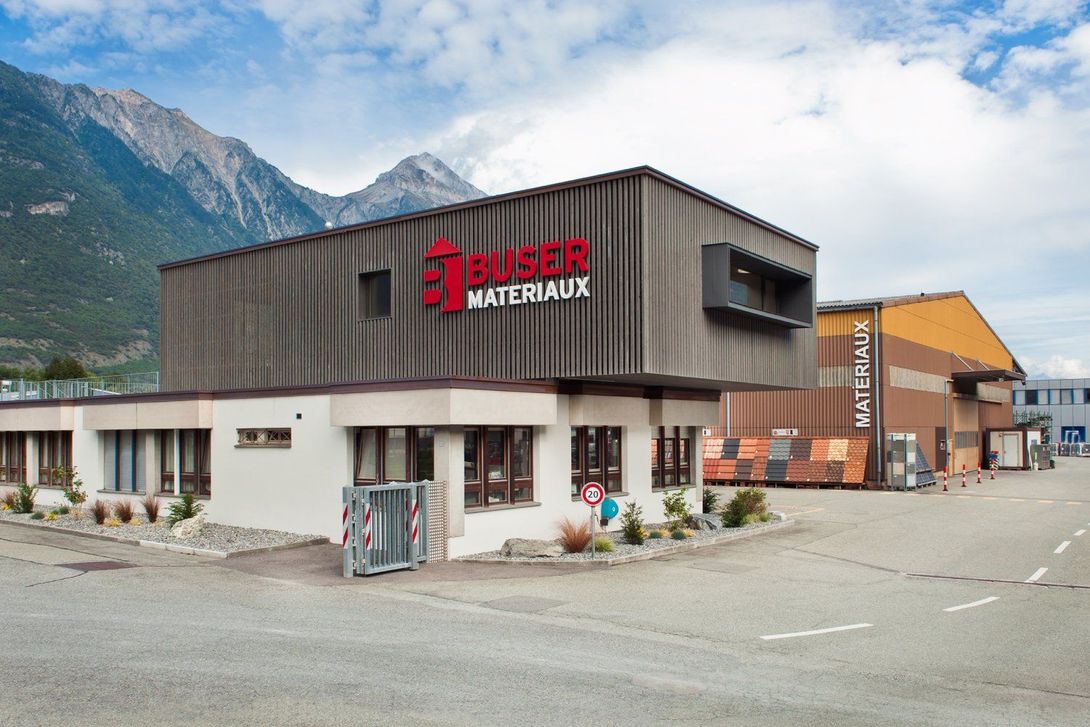 Un grand bâtiment avec des montagnes en arrière-plan et un panneau rouge sur le côté Buser Matériaux