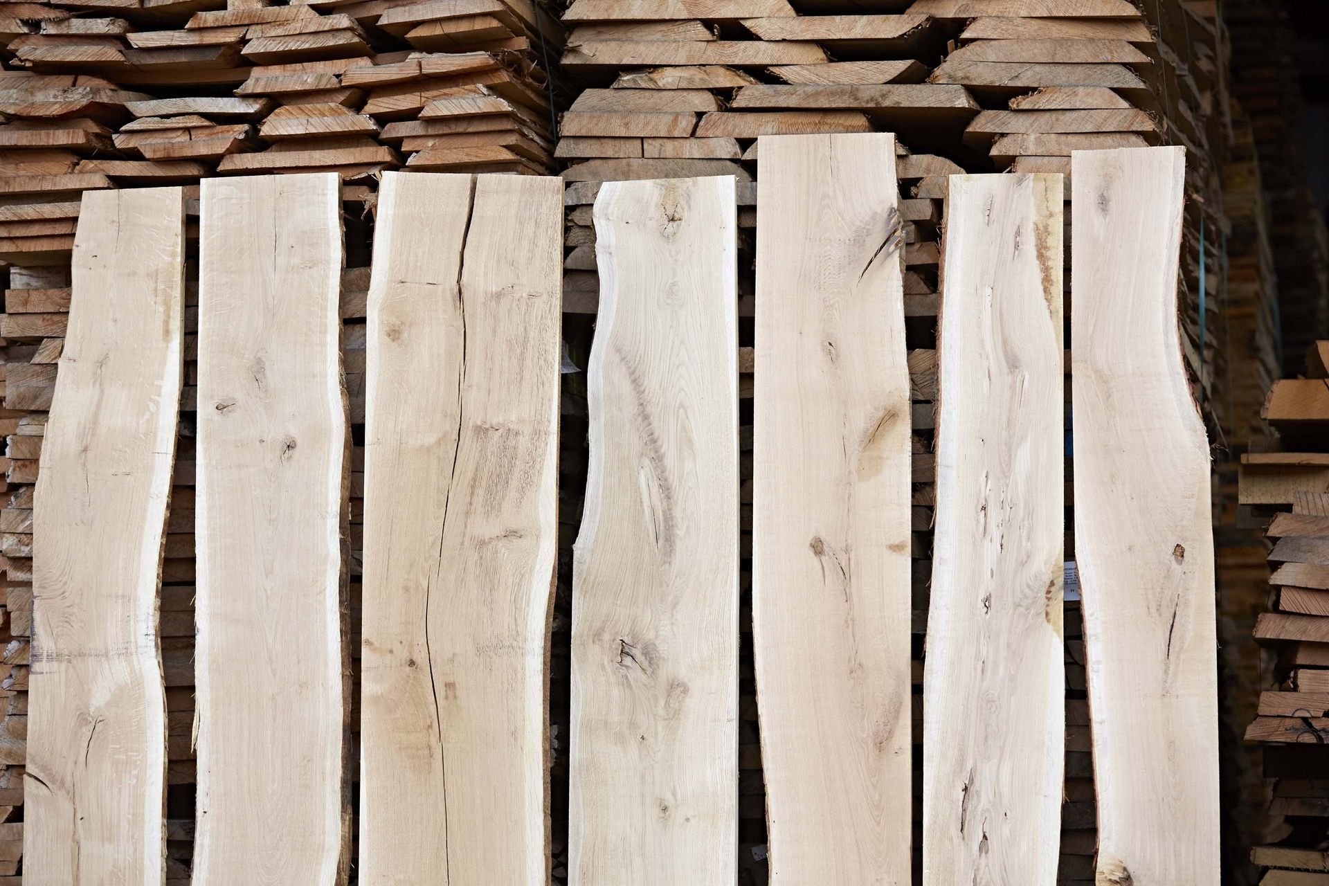 Sept planches de bois clair aux bords naturels, empilées devant d'autres morceaux de bois.