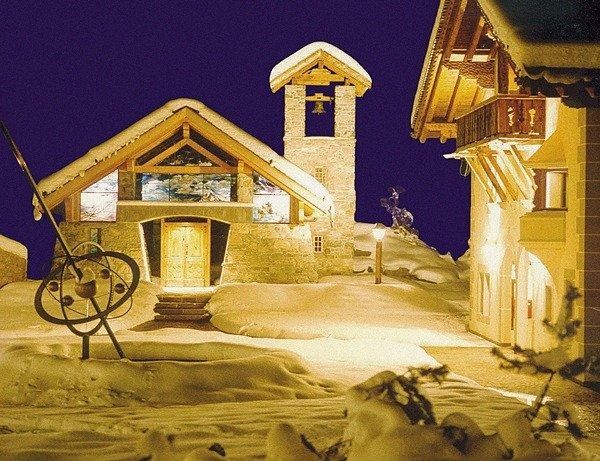 Scène de village enneigée la nuit, bâtiments en pierre aux intérieurs chaleureusement éclairés.