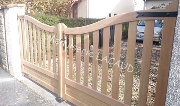 Portail d'allée en bois à lattes verticales et traverse supérieure incurvée, jouxtant un trottoir et un mur de briques.