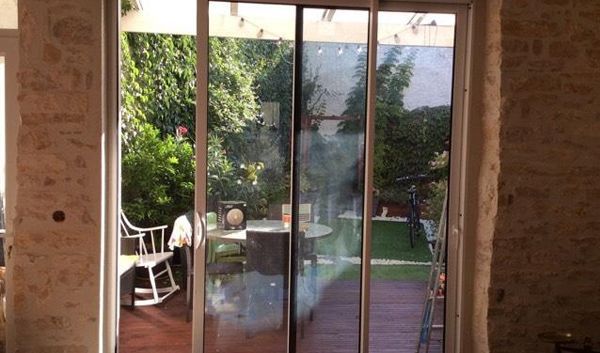Des portes-fenêtres s'ouvrent sur un jardin avec une table et des chaises sur une terrasse en bois.