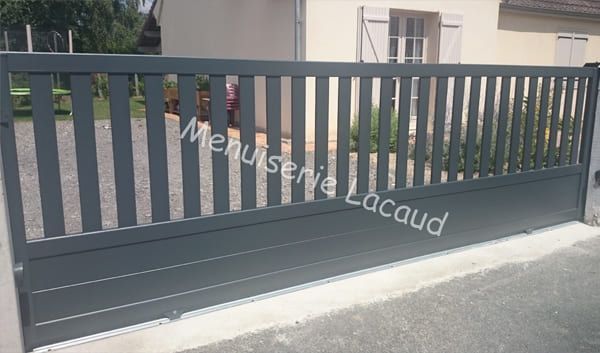 Portail coulissant en métal gris foncé devant un bâtiment beige, avec allée et trottoir.