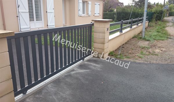 Portail d'entrée gris foncé à côté d'une maison beige, avec une allée pavée et un trottoir longeant un muret.