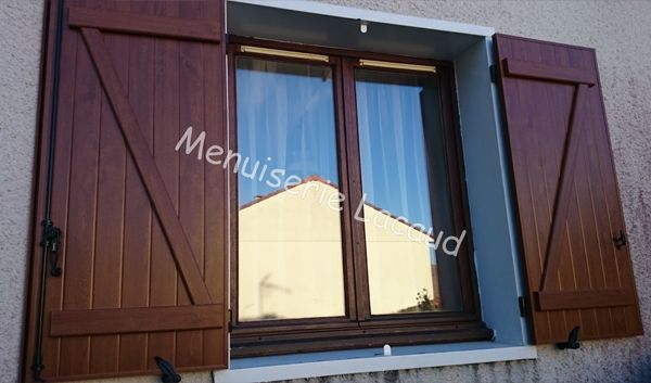 Fenêtre à volets bruns sur un mur de briques, avec reflet du ciel bleu et une toiture rouge.