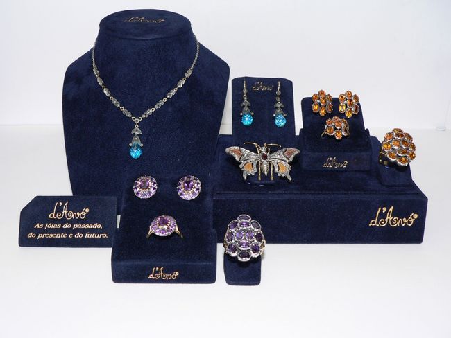 Expositor de joias sobre veludo azul: colares, brincos, anéis e um broche com pedras preciosas. Detalhes em dourado.
