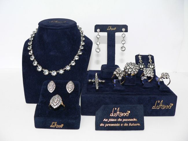 Expositor de joias sobre veludo azul: colar, brincos, anéis e um crucifixo. Texto dourado: 