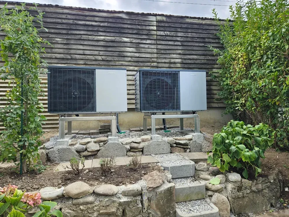 Zwei Wärmepumpen, die auf Metallgestellen in einem Garten an einem Holzzaun montiert sind.