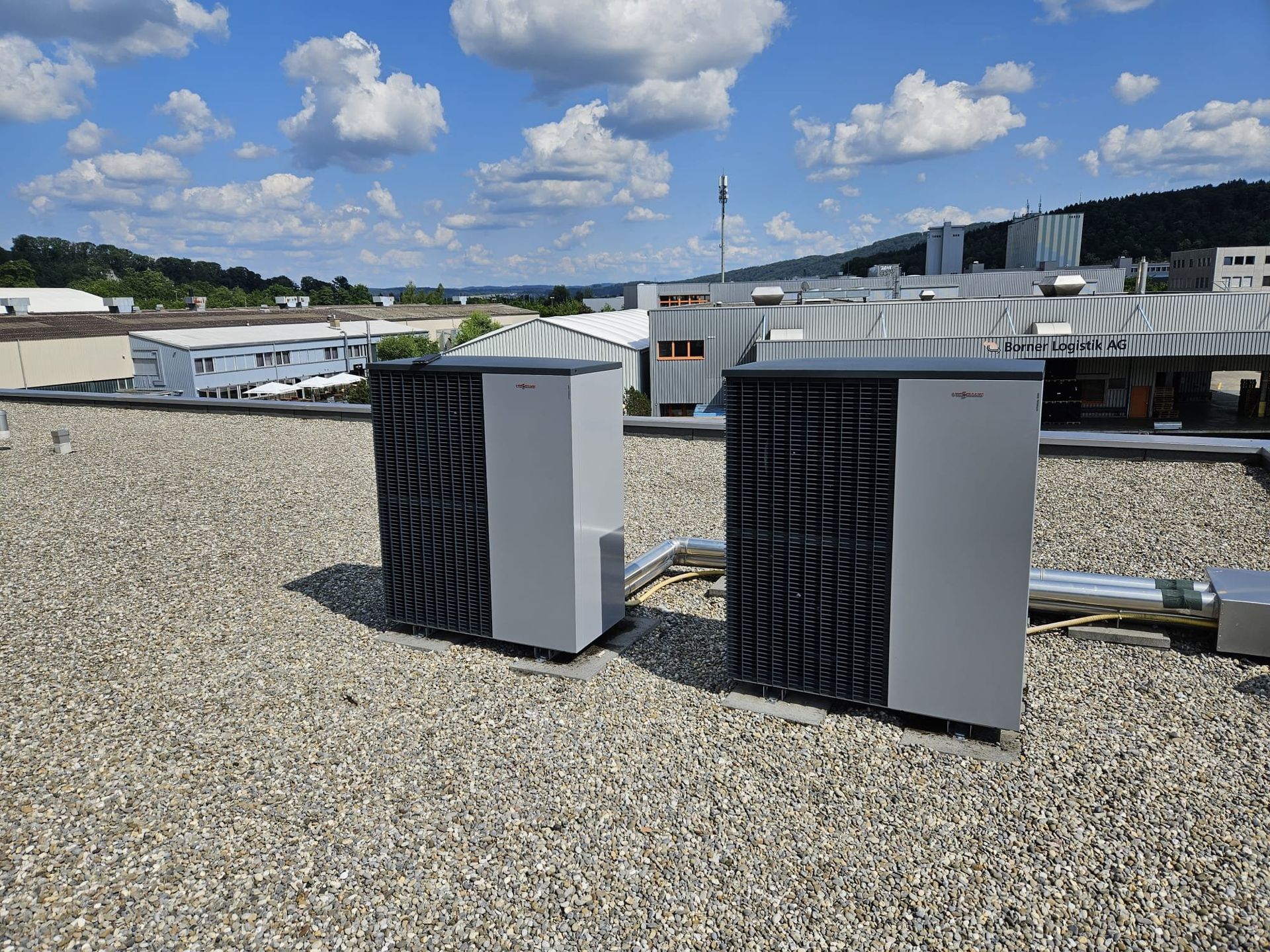 Zwei graue Klimaanlagen auf einem Kiesdach vor einem strahlend blauen Himmel mit Wolken.