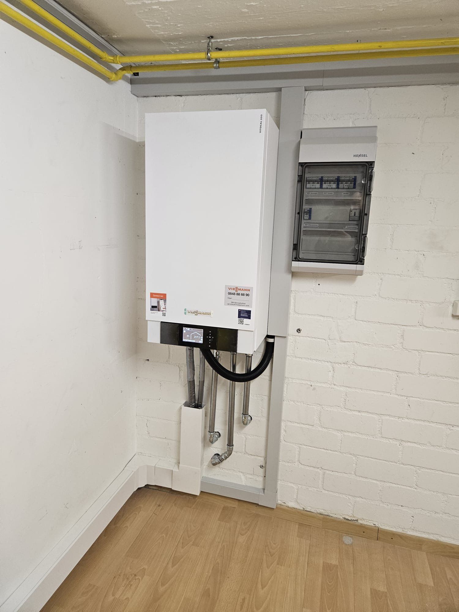 Ein weisser, an der Wand montierter Heizkessel in einer Zimmerecke, dazu silberne Rohre und ein grauer Elektrokasten.