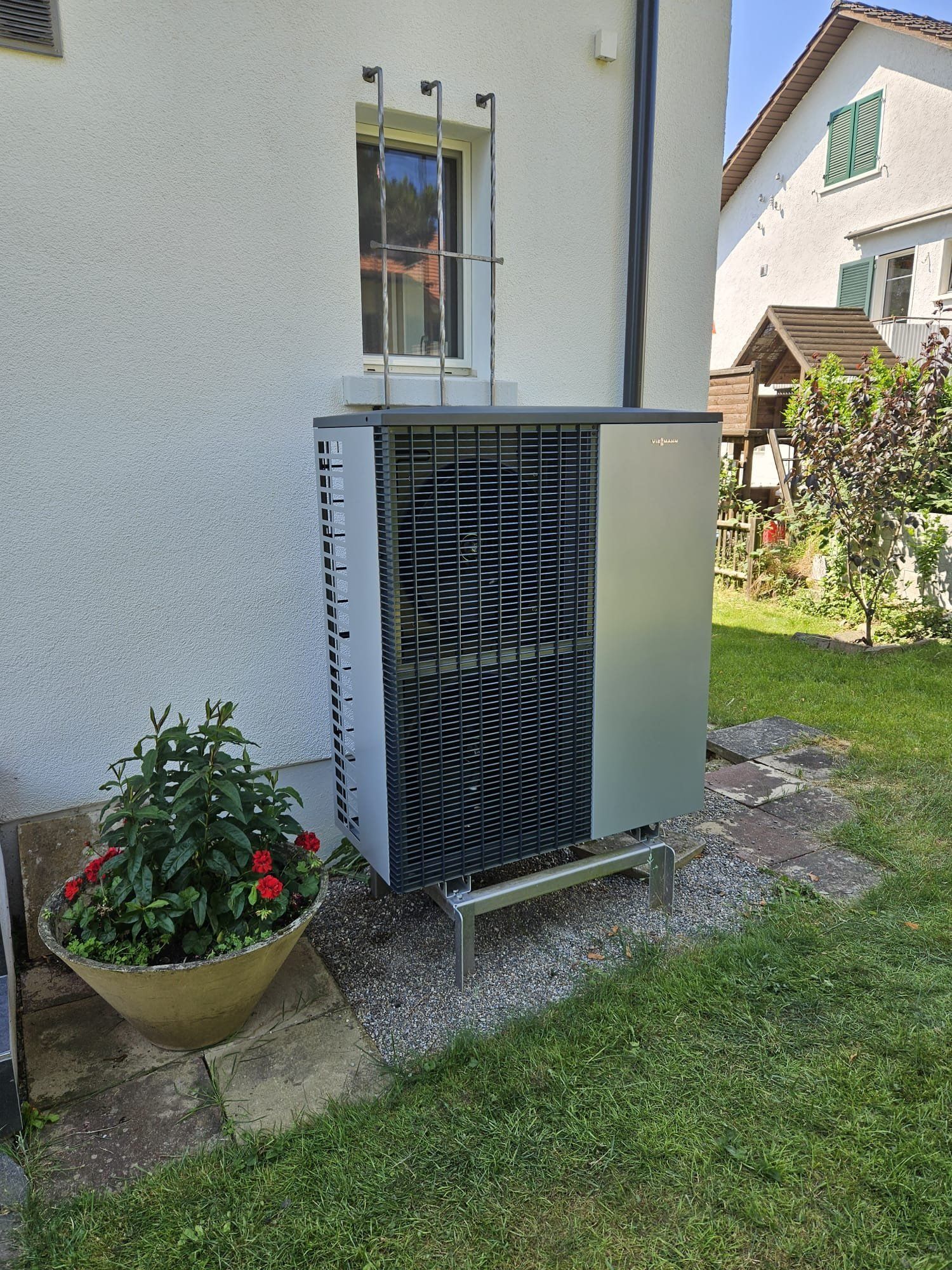 Eine Wärmepumpenanlage ist im Freien neben einem weissen Haus mit Fenster und einem daneben stehenden Blumentopf montiert.