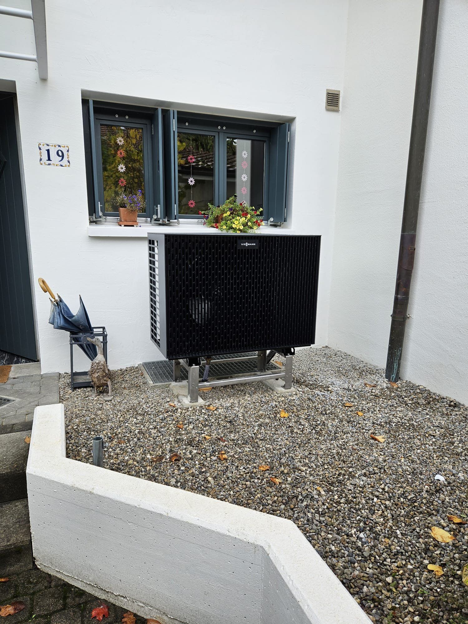 Schwarze Wärmepumpe ausserhalb eines weissen Gebäudes mit Fenster, auf Kies aufgestellt.