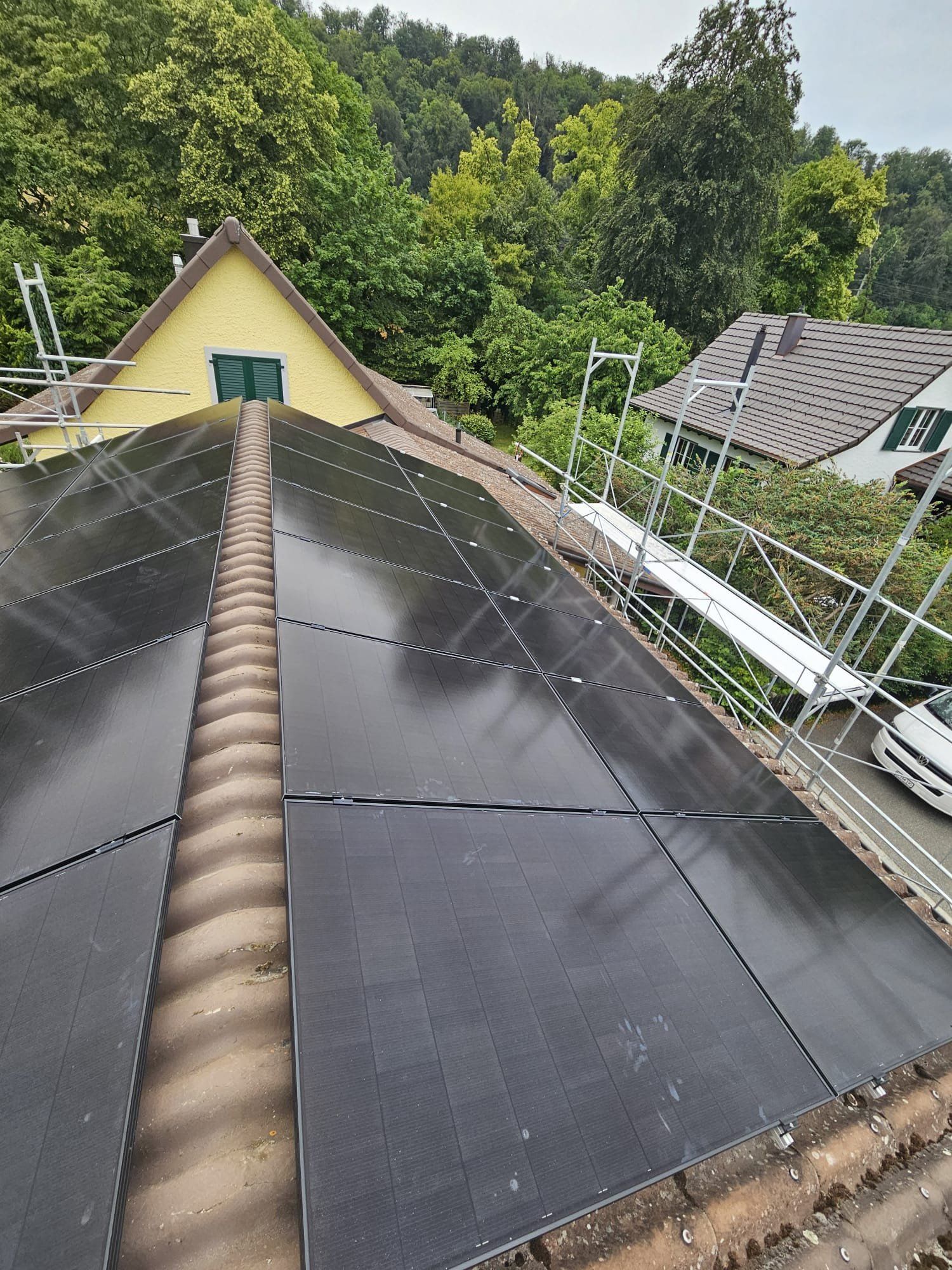Solarpaneele auf einem Dach, mit Gerüst. Bäume und ein Haus im Hintergrund.