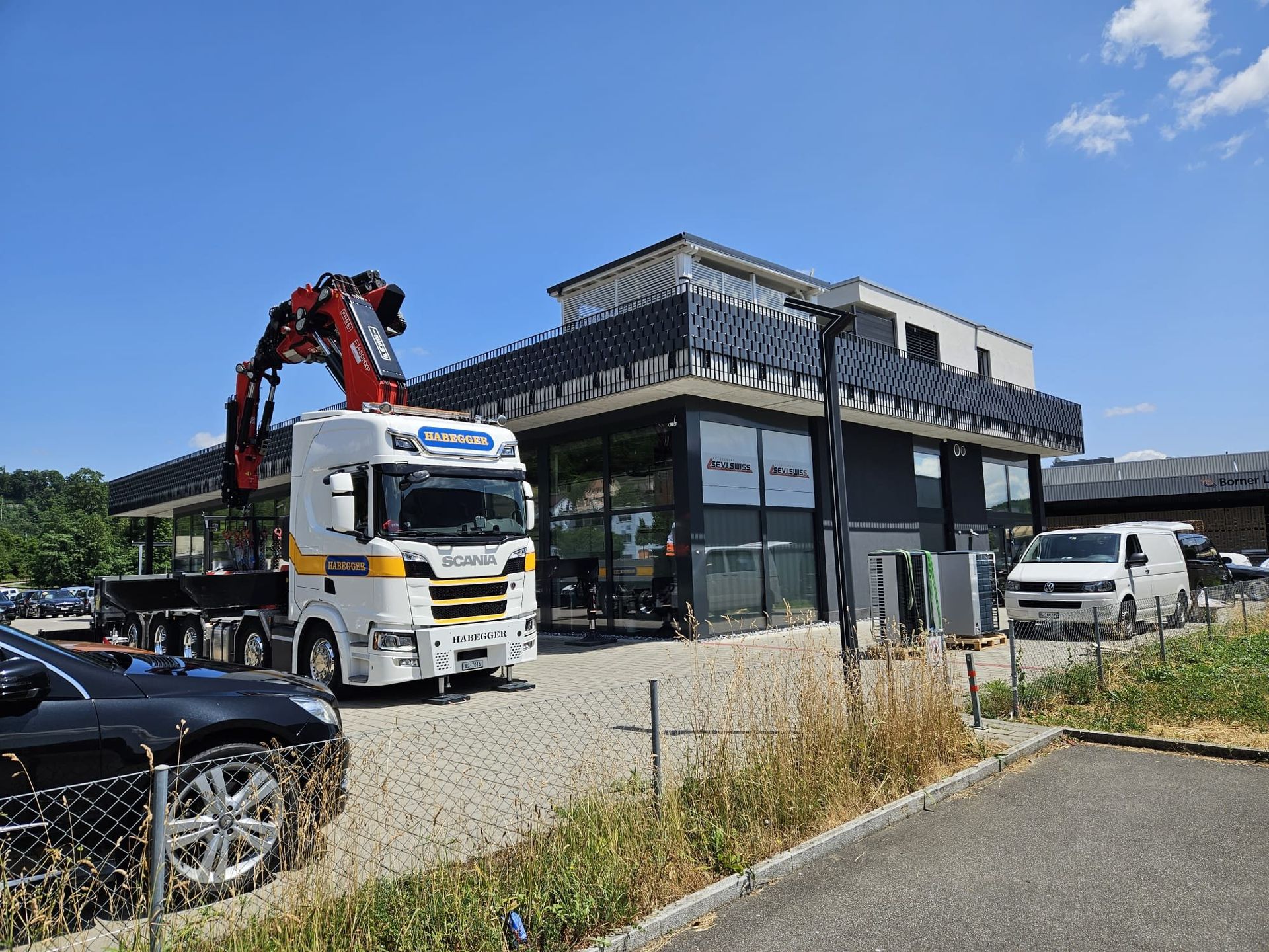 Ein LKW mit Kran vor einem modernen Gebäude an einem sonnigen Tag.