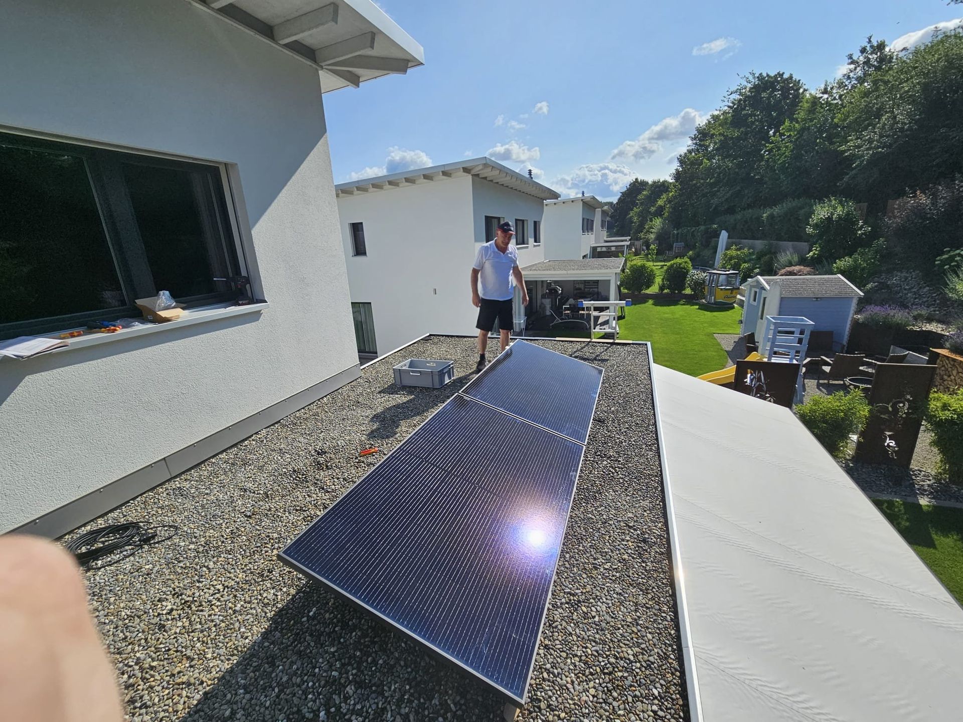 Mann steht an einem sonnigen Tag auf einem Kiesdach mit Solarpaneelen. Im Hintergrund sind ein Haus und Bäume zu sehen.