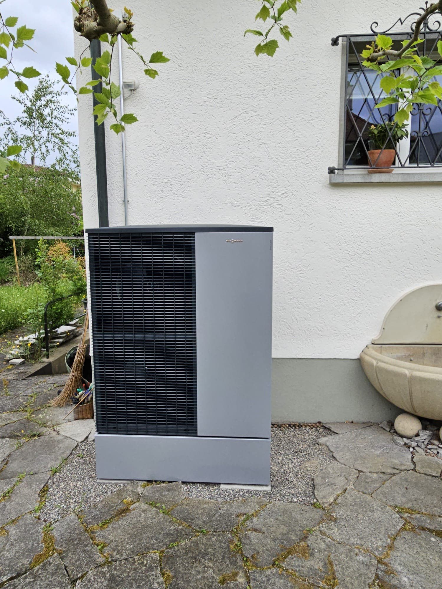 Eine graue Wärmepumpe vor einem weissen Haus neben einem kleinen Springbrunnen, darüber Laubwerk.