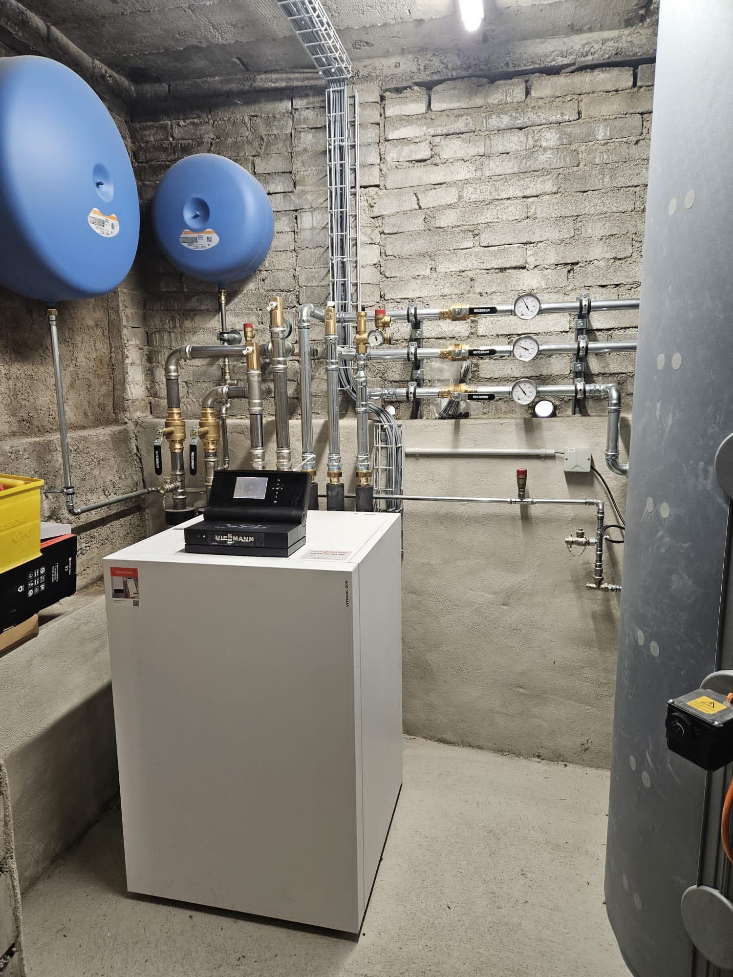 Ein Heizraum mit weissen Anlagen, blauen Tanks und verschiedenen Rohren und Messgeräten an einer Steinwand.