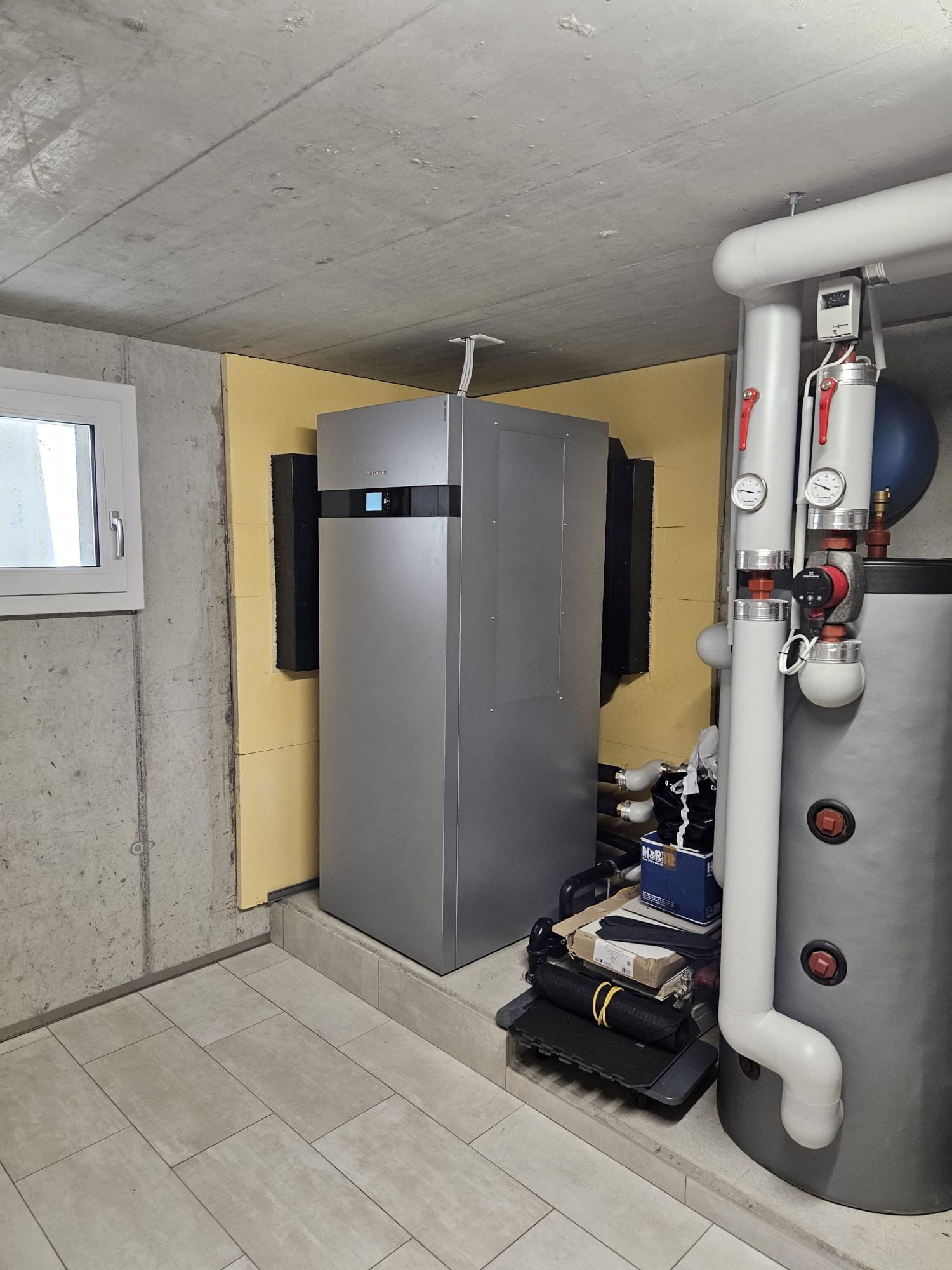 Eine silberne Wärmepumpe steht im Keller. Weisse Rohre und ein schwarzer Wassertank sind zu sehen.