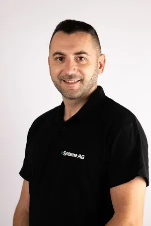 Ein Mann in schwarzem Hemd lächelt vor einem weissen Hintergrund. Auf dem Hemd ist das Logo der „Systeme AG“ zu sehen.