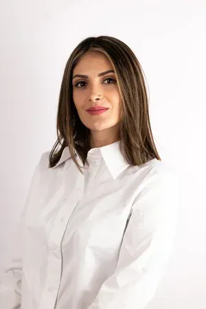 Frau mit braunen Haaren, die ein weisses Hemd mit Kragen trägt, schaut in die Kamera.