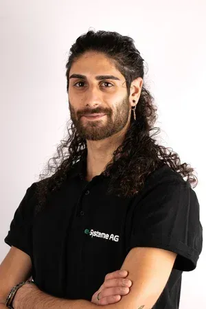 Mann mit lockigem braunem Haar, schwarzem Hemd, verschränkten Armen, lächelnd, neutraler Hintergrund.