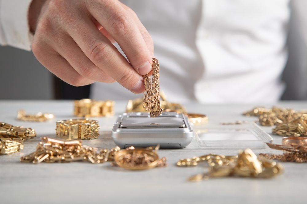 Una mano coloca joyas de oro sobre una báscula digital entre piezas de oro esparcidas sobre una mesa.