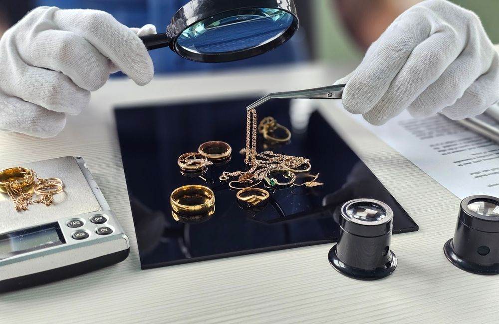 Unas manos enguantadas inspeccionan anillos de oro sobre una bandeja negra con una lupa y una regla.