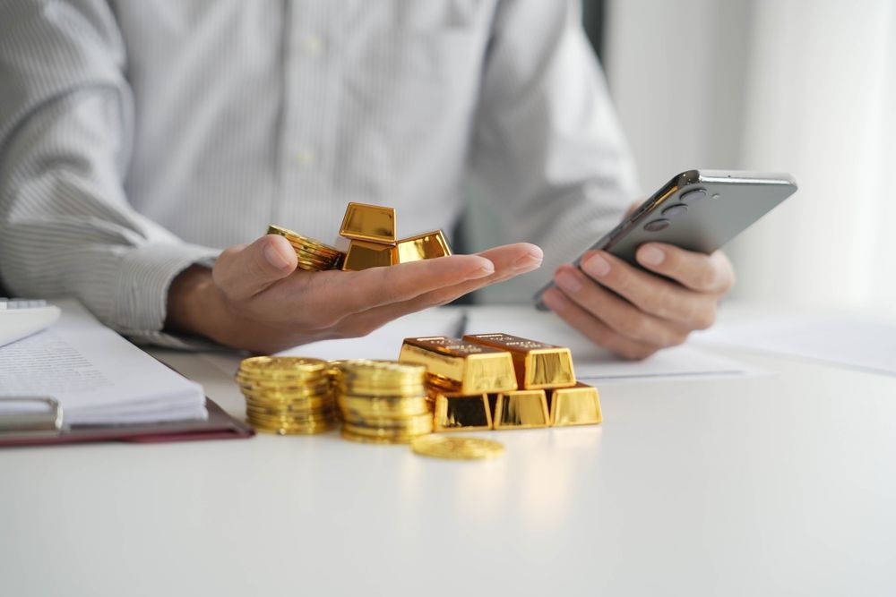 Persona sosteniendo un teléfono inteligente junto a pilas de lingotes de oro sobre un escritorio blanco.