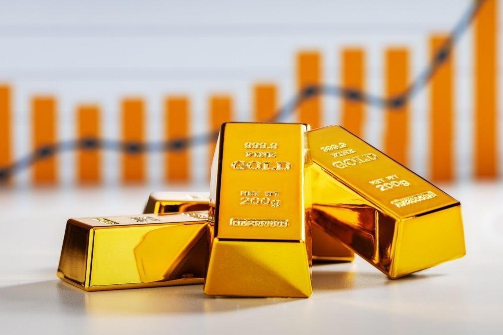 Lingotes de oro frente a un gráfico de barras naranja, lo que sugiere un alza en los mercados financieros.