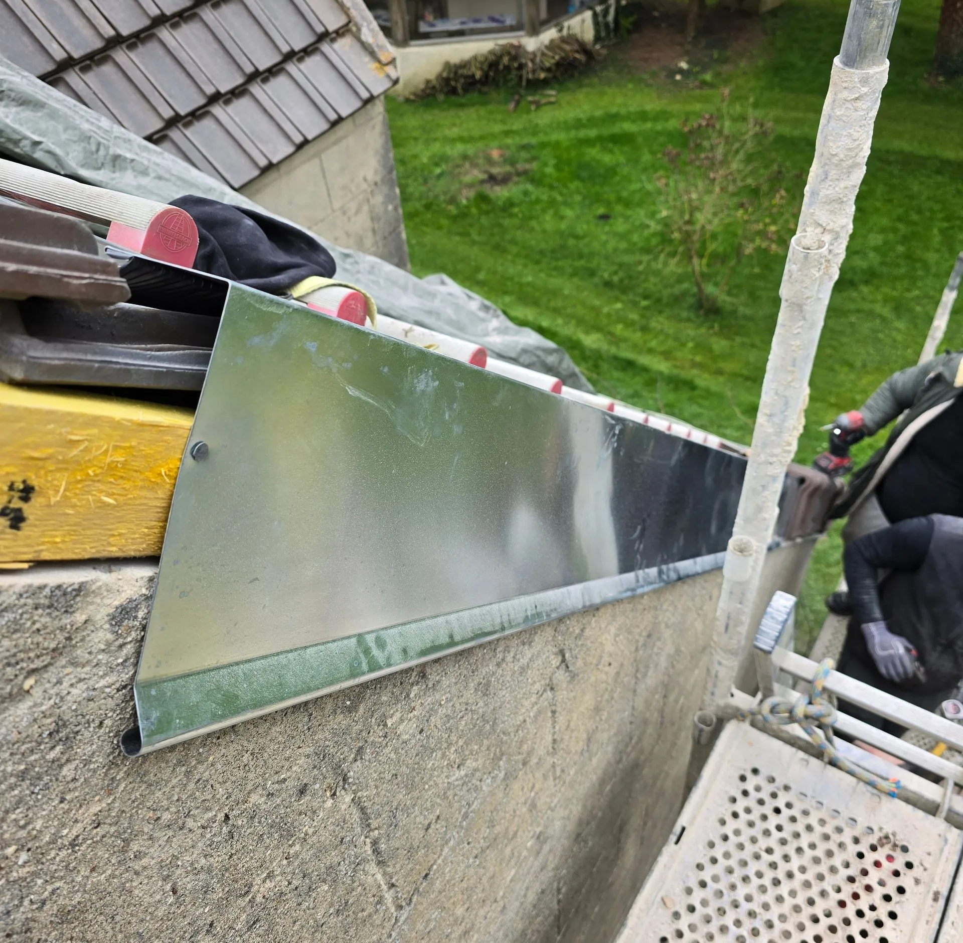 Des solins métalliques sont installés en bordure de toit, près d'un bâtiment et d'un échafaudage. Un ouvrier est également présent.