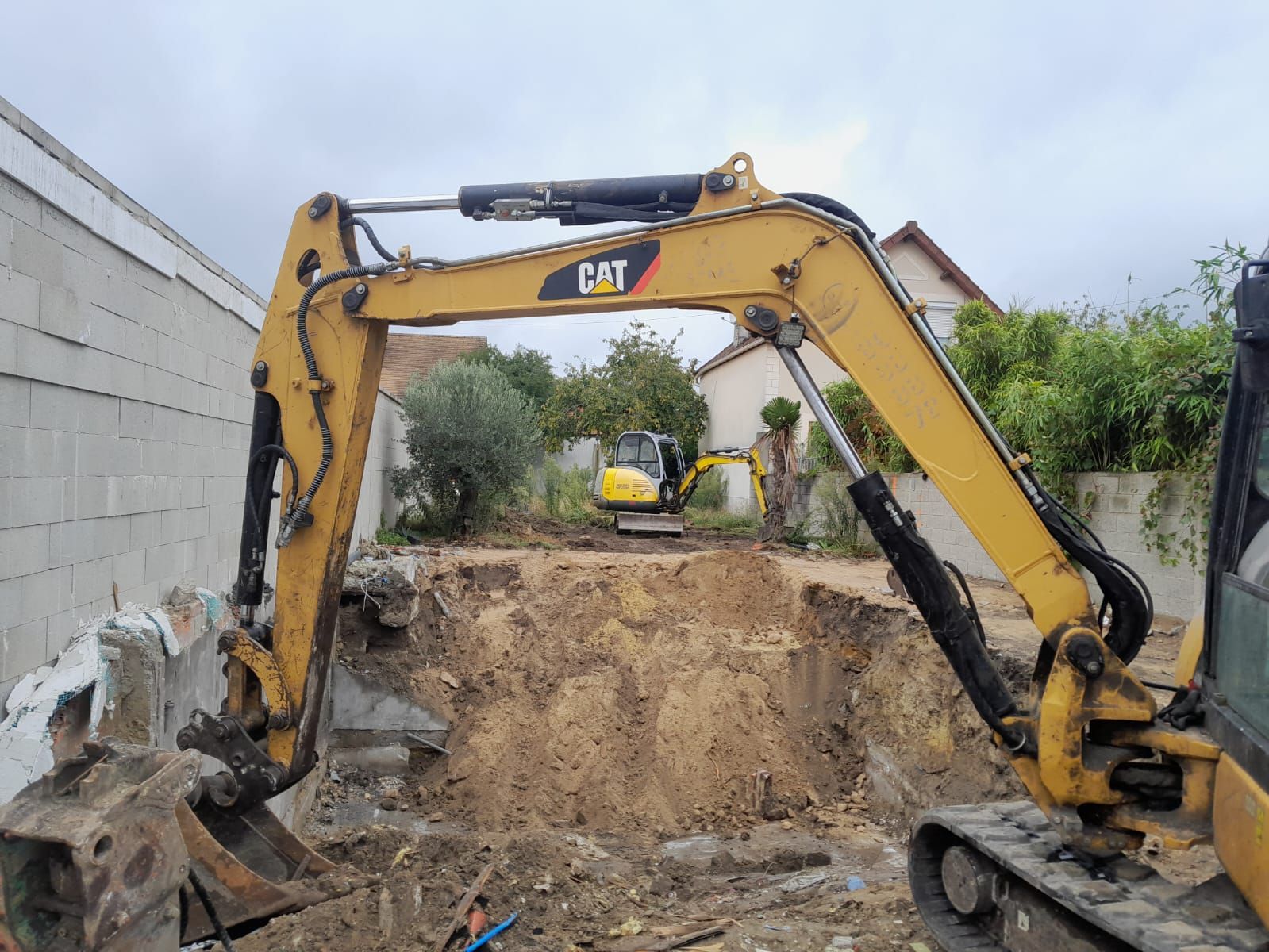 Une pelle mécanique jaune de type CAT creuse une fosse, une autre pelle mécanique est visible à l'arrière-plan. Chantier de construction.