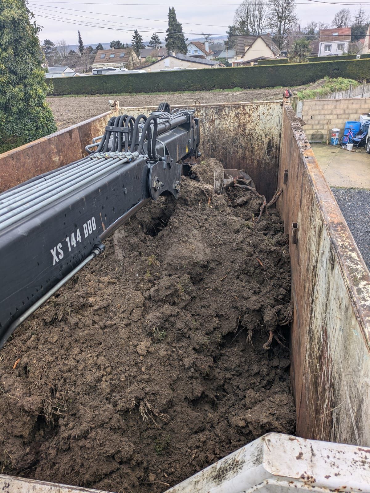 Le bras d'une excavatrice déverse de la terre dans un grand conteneur métallique, en extérieur.