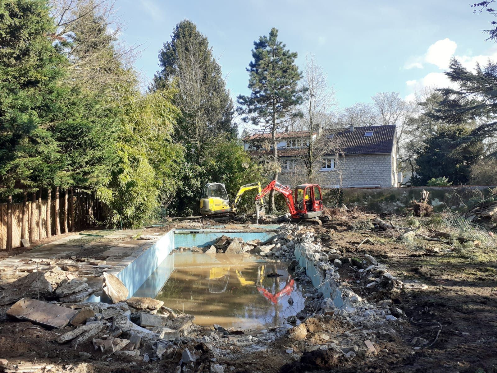 Chantier de démolition : des excavatrices dans un bassin, entourées de débris et d'arbres, par une journée ensoleillée.