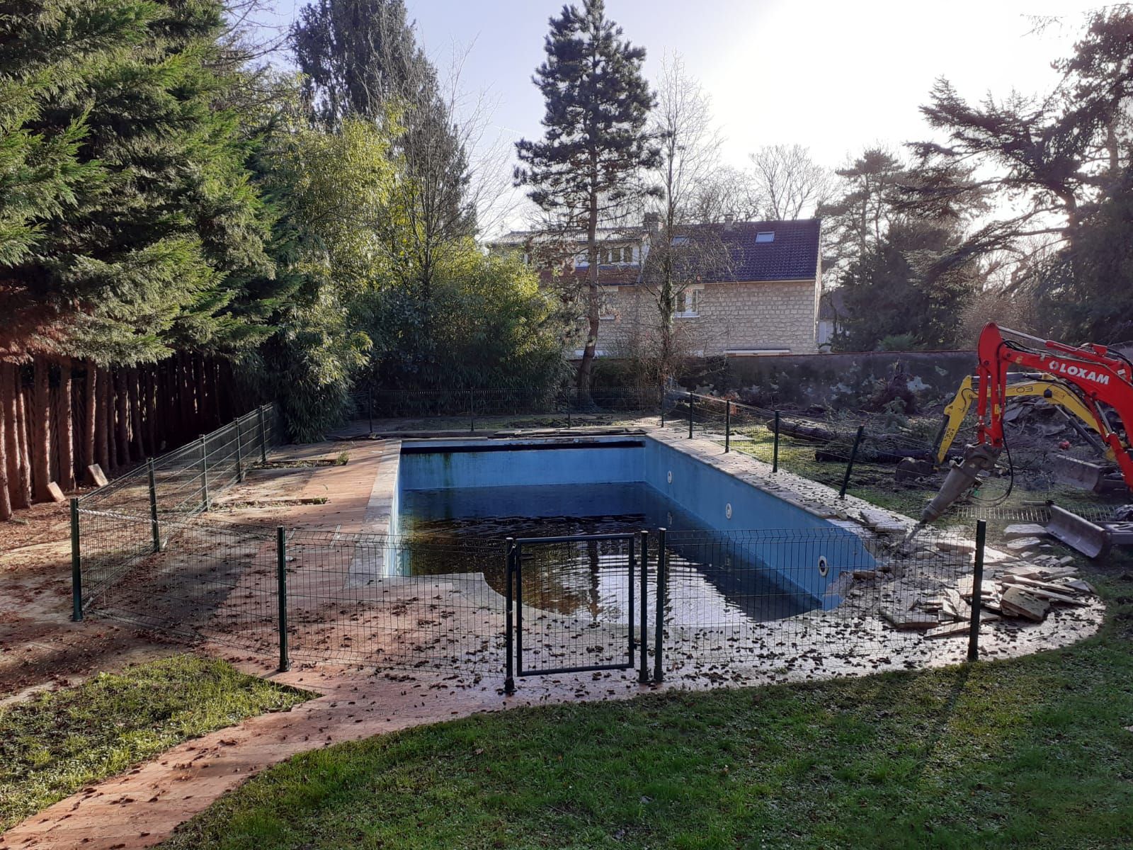 Piscine en cours de démolition, pleine d'eau, avec une excavatrice sur le côté.